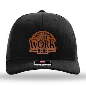 May include: Black trucker hat with a curved brim and mesh back. The front features a brown patch with the words "I DON'T KNOW I JUST WORK HERE" in a stylized font. A Richardson brand tag is visible.