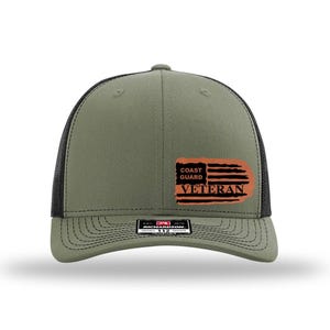 May include: Olive green and black trucker hat with a curved brim. The hat features a distressed orange patch with the words "Coast Guard Veteran" and a stylized American flag design. The hat has a mesh back.