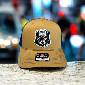 May include: A tan and navy blue Richardson 112 trucker hat. The hat features a patch of a raccoon wearing sunglasses and a hat with an American flag design. The hat is adjustable.