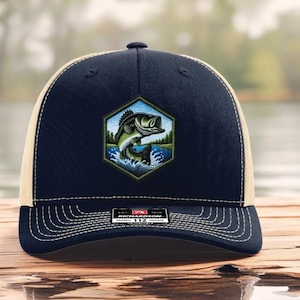May include: Navy blue and tan trucker hat featuring a hexagonal patch with a leaping bass illustration. The hat has a curved brim, mesh back, and a snap closure. The patch shows a green and black bass jumping out of the water.