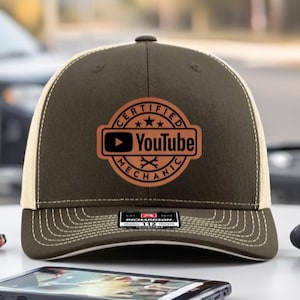 May include: A brown and beige trucker hat with the text "CERTIFIED YOUTUBE MECHANIC" in a circular logo. The hat is on a white surface with a phone, a charging cable, and a game controller.