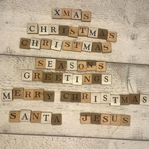 May include: Wooden letter tiles spelling out festive words including "XMAS", "CHRISTMAS", "SEASONS GREETINGS", "MERRY CHRISTMAS", "SANTA", and "JESUS". The tiles are arranged on a white wooden surface.