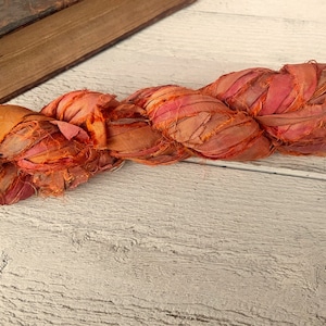 May include: A twisted strand of orange and pink sari silk ribbon. The ribbon is made of strips of fabric and has a textured appearance. The ribbon is lying on a white wooden surface, with a book in the background.