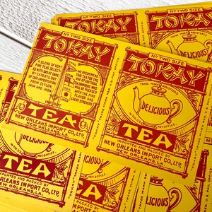 Puede incluir: Una colección de etiquetas de té Tokay vintage. Las etiquetas son amarillas con texto e ilustraciones rojas, incluyendo una tetera y las palabras "Delicious" y "Tea". Las etiquetas son de New Orleans Import Co., Ltd.