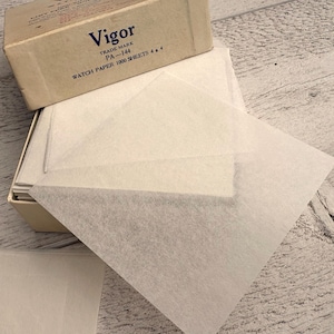 May include: A box of "Vigor" watch paper, PA-144, with 1000 sheets, each 4 x 4 inches. The box is beige with dark blue text. Several sheets of the watch paper are fanned out, showing their translucent quality.