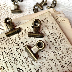 May include: A close-up of antique brass binder clips on a stack of old paper with handwritten text. The clips have a round top and a flat base.