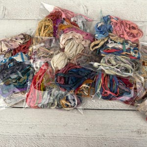 Könnte beinhalten: Sortiment bunter Seidenbandbündel in transparenten Plastiktüten. Die Bänder sind in verschiedenen Rosa-, Blau-, Rot- und Cremetönen gehalten. Die Bündel liegen auf einer weißen Holzoberfläche.