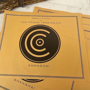 Puede incluir: Una etiqueta de papel marrón con un logotipo circular negro y el texto "Made in Finland for YUE FOONG TRADING CO. SHANGHAI" impreso en ella.