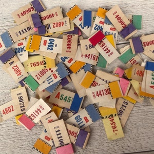May include: A pile of assorted vintage paper tags in shades of yellow, pink, blue, and purple. Each tag features printed text and numbers in red and black. The tags are arranged on a light-coloured wooden surface.