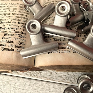 Könnte beinhalten: Nahaufnahme eines Stapels silberner Büroklammern auf einem geöffneten Buch. Das Buch ist alt und hat vergilbte Seiten mit schwarzem Text. Die Büroklammern sind aus Metall und haben eine glatte, glänzende Oberfläche.