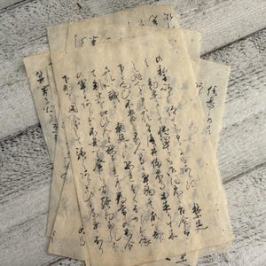 Puede incluir: Una pila de hojas de papel envejecido de color crema con caligrafía manuscrita en tinta negra. El papel tiene una superficie ligeramente texturizada y parece antiguo. La caligrafía es intrincada y detallada, con una escritura tradicional de Asia Oriental.