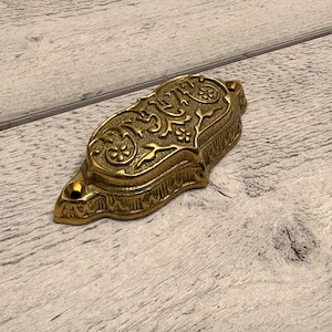 Könnte beinhalten: Ein goldfarbener, verzierter Metall-Schubladengriff mit einem komplizierten floralen Design. Der Griff ist rechteckig mit einer gewölbten Oberseite und einem kleinen Loch zur Befestigung. Die Oberfläche hat ein strukturiertes, antikes Aussehen.