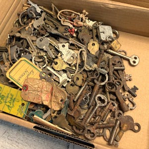 Könnte beinhalten: Eine Pappschachtel voller antiker Schlüssel. Die Schlüssel variieren in Größe, Form und Metallart, einige weisen Alters- und Gebrauchsspuren auf. Einige Anhänger sind sichtbar, darunter einer mit der Aufschrift "Kent House."