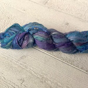 K&ouml;nnte beinhalten: Ein Kn&auml;uel aus blauem und violettem Seidenband, verdreht und verknotet, auf einer wei&szlig;en Holzoberfl&auml;che.