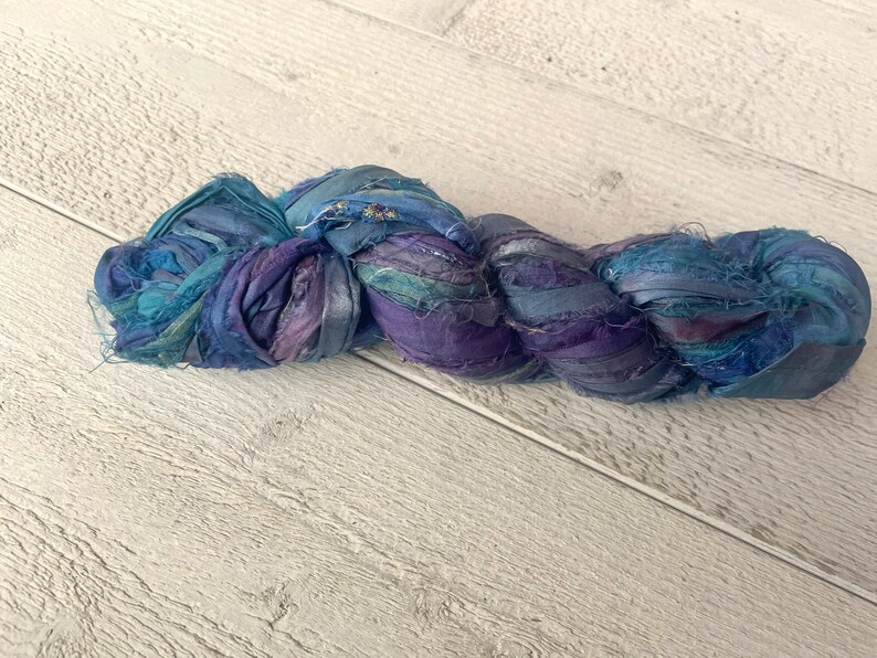 K&ouml;nnte beinhalten: Ein Kn&auml;uel aus blauem und violettem Seidenband, verdreht und verknotet, auf einer wei&szlig;en Holzoberfl&auml;che.