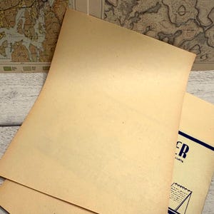 Op de afbeelding: Verschillende vellen verouderd, crèmekleurig papier zijn opgestapeld, met een hoek van een bedrukte pagina zichtbaar. Het papier heeft een licht getextureerd oppervlak en een vintage esthetiek. De achtergrond bevat delen van antieke kaarten.