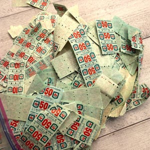Op de afbeelding: Een stapel vintage S&H Green Stamps, met het nummer "50" in rood en de woorden "Sperry and Hutchinson" in een groen en wit ontwerp. Sommige postzegels tonen het nummer "10". De postzegels liggen op een lichte achtergrond.