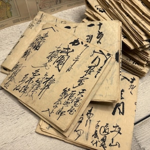 Può includere: Una pila di quadrati di carta invecchiata con calligrafia nera. La carta ha una tonalità marrone chiaro e l'inchiostro sembra essere nero. La calligrafia è in stile tradizionale dell'Asia orientale. L'immagine mostra una collezione di documenti d'epoca.