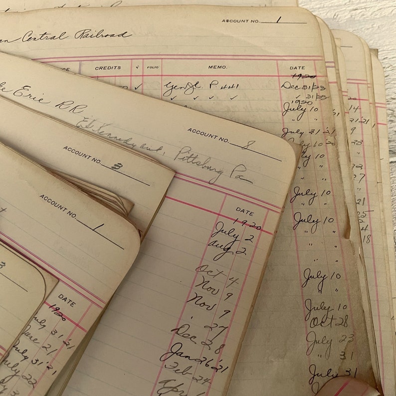 May include: Close-up of a stack of vintage ledger books with handwritten entries and dates. The books are labeled with account numbers and have pink lines. The text on the pages includes "Central Railroad", "Erie RR", "General P.I.", and "Rent, Pittsburg Pa."