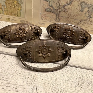 Pode incluir: Três puxadores de gaveta antigos em bronze com um design floral. Cada puxador tem uma placa traseira oval com uma flor central e volutas. Os puxadores estão sobre uma superfície de madeira clara, com mapas antigos no fundo.