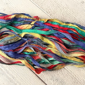 Pode incluir: Um monte de sobras de fita de seda colorida em tons de azul, vermelho, amarelo e verde. A fita está disposta em um monte solto e fluido sobre uma superfície de madeira branca.