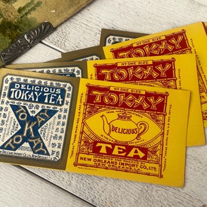 Puede incluir: Etiquetas de té Tokay vintage en amarillo y dorado. Las etiquetas presentan el texto "Delicious Tokay Tea" en rojo y azul, con una ilustración de tetera. Las etiquetas son de New Orleans Import Co.