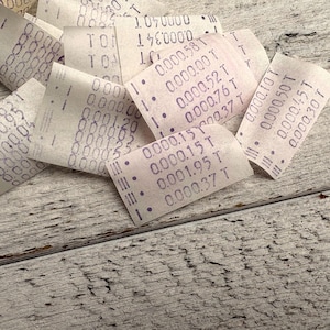 May include: A close-up of several small, curled paper receipts with printed text in purple ink. The receipts are scattered on a weathered, light-coloured wooden surface. The text includes numbers and the letter "T".