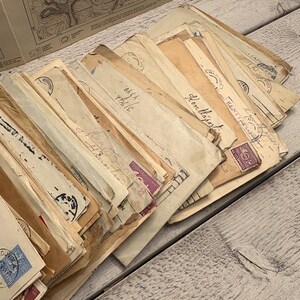 May include: A collection of vintage envelopes, varying in size and color, with stamps and handwritten text. The envelopes are arranged in a pile, showcasing their aged appearance. Some envelopes have purple stamps and blue stamps.