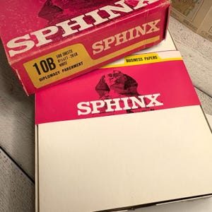 Könnte beinhalten: Zwei Kartons Geschäftspapier der Marke Sphinx. Die obere Box ist rot mit weißem Text und einem gelben Etikett mit der Aufschrift "10B" und "500 Blatt 21,6 x 27,9 cm - 9,07 kg White Diplomacy Parchment". Die untere Box ist weiß mit einem roten Band und dem Sphinx-Logo.