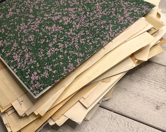 Puede incluir: Una pila de documentos de papel envejecido y un cuaderno. El cuaderno tiene una cubierta verde con un patrón moteado rosa. Los papeles son de varios tamaños y tonos de papel amarillento, algunos con texto manuscrito. Los artículos están sobre una superficie de madera clara.