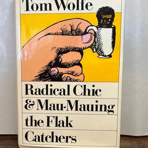 May include: A paperback book titled "Radical Chic & Mau-Mauing the Flak Catchers" by Tom Wolfe. The cover features a hand holding a mug with a person's face inside, set against a yellow background.