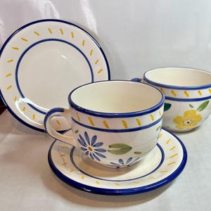 May include: A white ceramic teacup and saucer set with blue and yellow floral designs. The set includes a teacup, saucer, and a larger plate, all with blue rims and decorative accents. The teacup has a handle and is decorated with blue flowers.