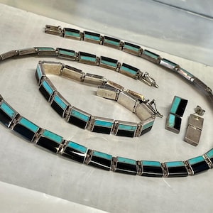 Op de afbeelding: Een zilverkleurige sieradenset met een ketting, armband en oorbellen. Elk stuk is versierd met afwisselend zwarte en turquoise rechthoekige inlays. De set wordt gepresenteerd op een lichtgekleurd oppervlak.