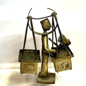 May include: A brass sculpture of a vendor carrying goods on a shoulder pole. The vendor figure is in profile, wearing a hat, and holding a small object. Two rectangular containers hang from the pole, each with a decorative floral design.
