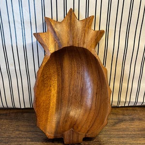 May include: A wooden pineapple-shaped bowl with a natural wood grain finish. The bowl has a carved design with a textured exterior and a smooth interior. The bowl is designed to resemble a pineapple fruit.