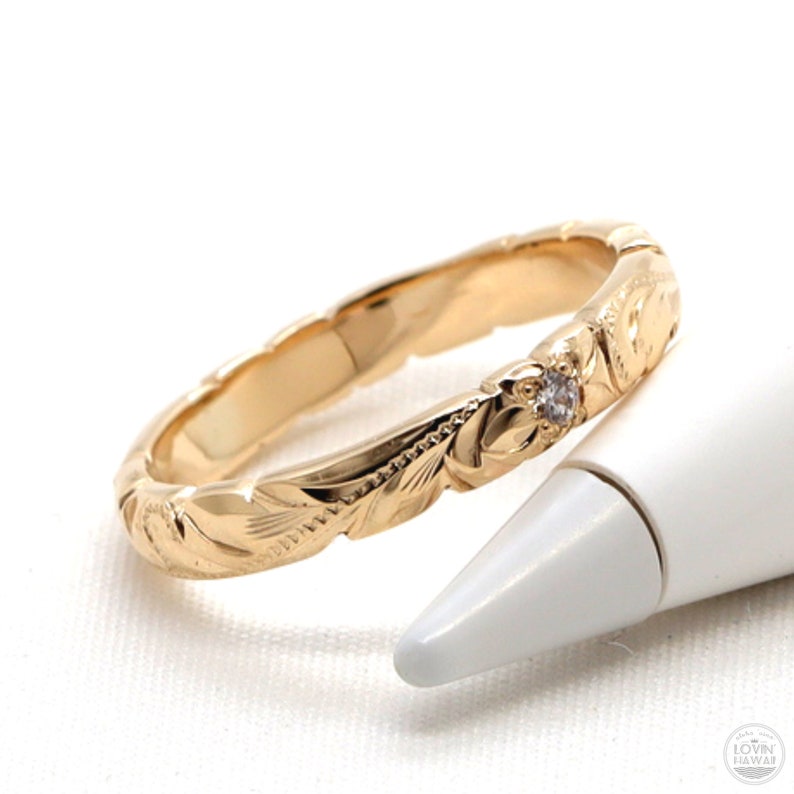 Puede incluir: Un anillo de banda de oro con un dise&ntilde;o floral y un solo diamante. El anillo est&aacute; sobre una superficie blanca.