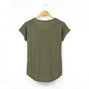 Rear view of olive green women’s t-shirt made from breathable slub cotton, displayed on a wooden hanger. The scoop neck and rolled sleeves offer a relaxed, feminine fit, while the dropped hem provides a flattering silhouette. This sustainable casual top is ideal for layering in a laid-back boho or earthy wardrobe.