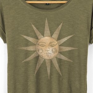 Close-up of the vintage sun face graphic on an olive women’s T-shirt, showing fine line details and soft tan ink, lightweight slub cotton texture visible around the scoop neckline.
