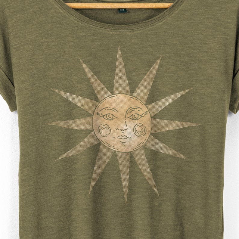 Close-up of the vintage sun face graphic on an olive women’s T-shirt, showing fine line details and soft tan ink, lightweight slub cotton texture visible around the scoop neckline.