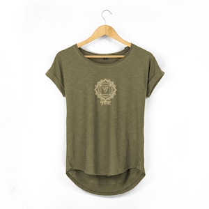May include: Olive green t-shirt with a crew neck and rolled sleeves. The shirt features a gold-colored geometric design with text in the center. The shirt is hanging on a wooden hanger against a white background.