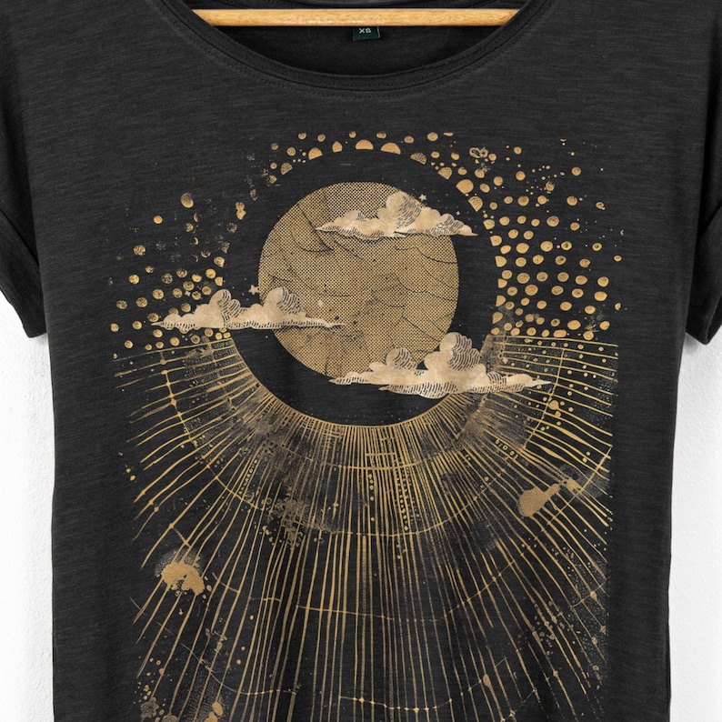 Close up of celestial moon graphic on black t shirt showing detailed clouds, circular moon design and radiating lines in a soft distressed gold print