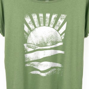 May include: Green t-shirt featuring a white graphic of a sun setting over the ocean. The sun has textured rays, and the waves are depicted with horizontal lines. The shirt is made of a soft material and has a relaxed fit.