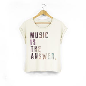 May include: A cream-coloured short-sleeve t-shirt with rolled sleeves and a round neckline. The front features the text "MUSIC IS THE ANSWER." printed in a dark, multi-coloured, textured graphic pattern that resembles a starry night sky.