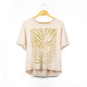Puede incluir: Camiseta beige claro con un diseño de hojas doradas. La camiseta de manga corta está colgada en una percha de madera sobre un fondo blanco. El diseño de hojas está centrado en la parte delantera de la camiseta.