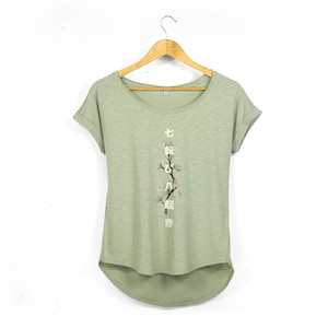 Peut inclure: T-shirt vert clair à col rond et manches retroussées. Le t-shirt présente un motif floral et des caractères japonais imprimés verticalement. Il est suspendu à un cintre en bois sur fond blanc.