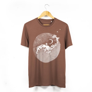 May include: A brown t-shirt with a white and light brown circular graphic. The graphic depicts a wave, a crescent moon, and a spiral design. The t-shirt is suspended on a wooden hanger against a white backdrop.