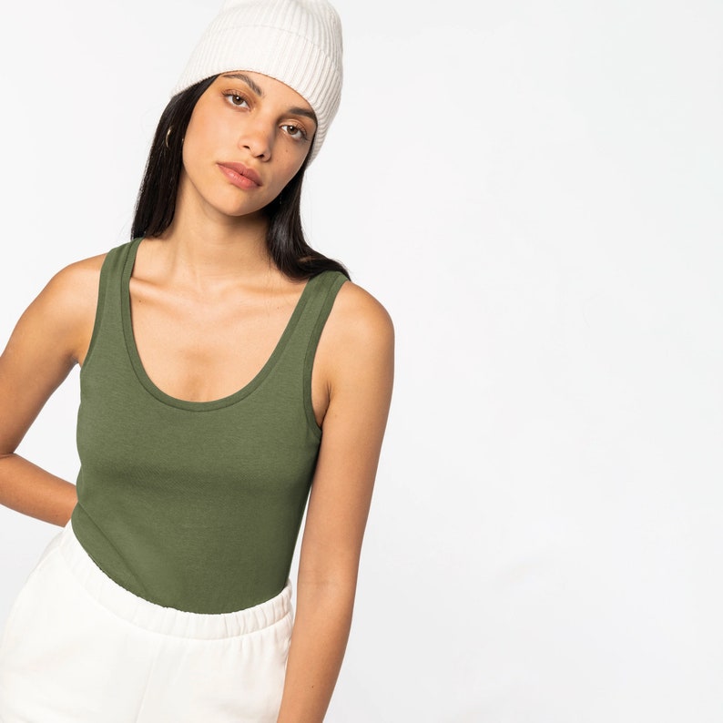 May include: A woman wearing a green tank top and a white beanie. She is wearing white trousers and has long dark hair.