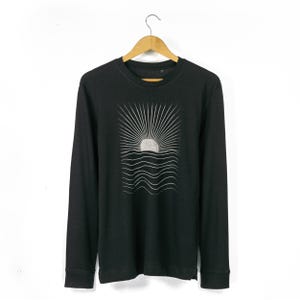 Puede incluir: Camiseta negra de manga larga con un gráfico blanco de un sol saliendo sobre líneas onduladas.