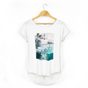 White relaxed fit women's t-shirt with rolled sleeves featuring a coastal collage graphic made from ocean waves, tropical palms, a surfboard silhouette, seaside horizon and surfer photography in muted turquoise, seafoam and soft grey tones.