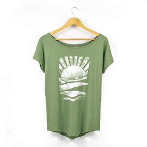 May include: Olive green t-shirt with a white graphic of a sun setting over rolling hills. The shirt has a scoop neck and short sleeves, hanging on a wooden hanger against a white background. A casual, nature-themed top.